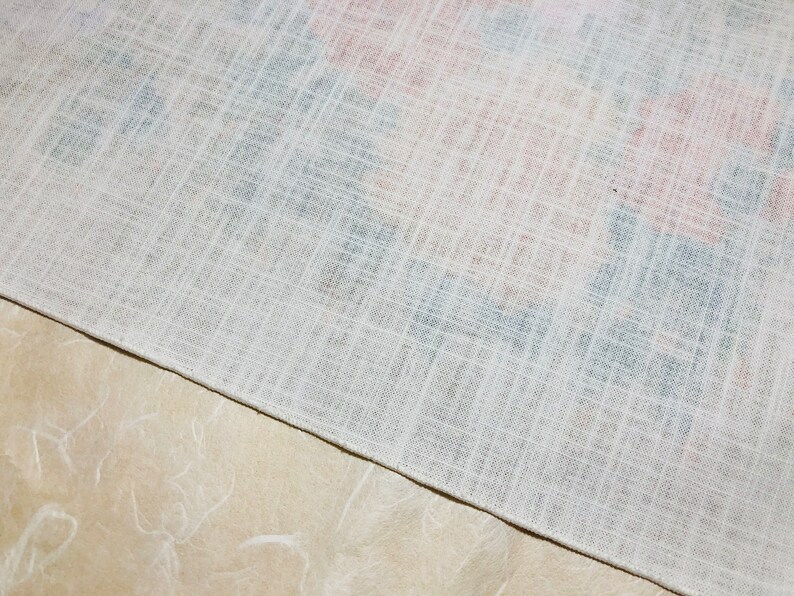 May include: Close-up of a white linen fabric with a subtle crosshatch pattern. Hints of red and blue floral designs are visible beneath the surface. The fabric is folded over a light beige surface.
