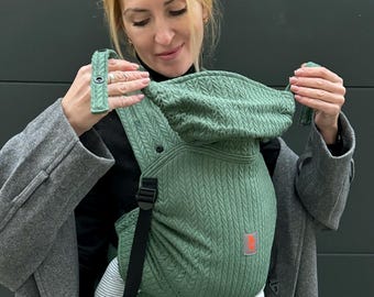 Puede incluir: Una mujer lleva un portabebés verde con un patrón de punto de cable. El portabebés tiene una hebilla negra y un logotipo rojo. La mujer lleva un abrigo gris y pantalones negros.