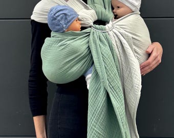 May include: A baby carrier in sage green and light gray holds two babies, each wearing a hat. The carrier is wrapped around the person's torso and secured with a metal ring. The person is wearing a black top and pants.