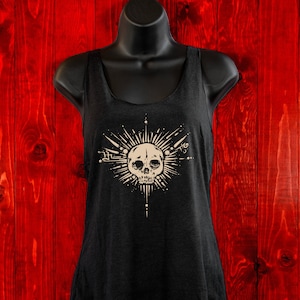 May include: A black tank top with a tan skull and starburst design. The design features a skull with a starburst of rays around it. The tank top is on a mannequin against a red wood background.