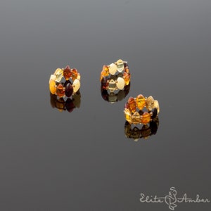 May include: Three amber rings with different color variations. The rings are made of small, round amber beads in shades of yellow, orange, brown, and red. The rings are arranged on a reflective surface.