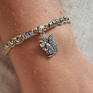 May include: A macrame bracelet with a silver owl charm. The bracelet features a tan cord with silver beads and decorative metal accents. The owl charm is detailed with etched feathers and eyes. The bracelet is worn on a wrist.