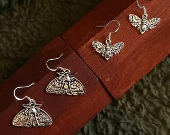 May include: Silver moth earrings on a dark wooden surface. The earrings have detailed wing patterns and curved ear wires. The background is a textured, dark green fabric. The earrings are a unique accessory.