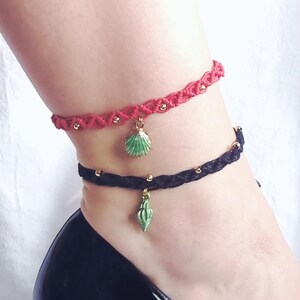 May include: Two braided anklets, one red and one black, each adorned with gold beads and a seashell charm. The anklets are worn on an ankle, paired with a black high-heeled shoe. The seashell charms are green.