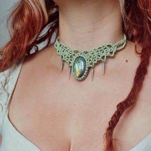 May include: A green macrame necklace with a central oval stone displaying iridescent colours. The necklace has silver-coloured spikes hanging below the stone and macrame design. The intricate knotwork creates a unique piece of jewellery. A statement necklace.