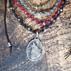 Può includere: Una collezione di collane in macramè fatte a mano in vari colori, tra cui nero, rosso, verde e blu. Ogni collana presenta un design a cordoncino intrecciato con perline color argento e un ciondolo con un lupo che ulula alla luna.
