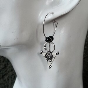 May include: Silver-toned hoop earring with a geometric pendant. The pendant features a triangle shape with a circular design in the center and small beaded accents. Black beads are at the top of the earring.