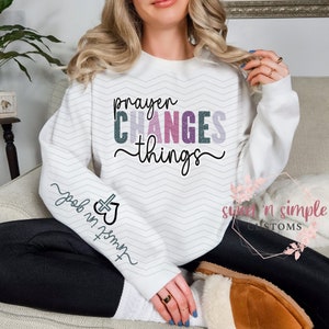 Peut inclure: Sweat-shirt blanc avec le texte "prayer CHANGES things" en lettres pailletées roses, violettes et bleues. La manche gauche porte le texte "trust in god" avec un graphique de cœur et de croix.