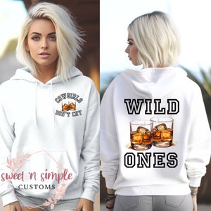 May include: White hoodie with a black and white graphic on the front that says "Cowgirls Don't Cry" and two glasses of whiskey. The back of the hoodie has a black and white graphic that says "Wild Ones" with two glasses of whiskey.