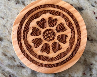 May include: Circular wooden coaster with a laser-etched floral design. The coaster features a central flower-like motif surrounded by intricate geometric patterns. The wood has a light, natural colour, and the etched design is a darker brown.