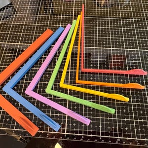May include: A collection of colorful, L-shaped tools in shades of orange, blue, purple, green, and yellow. These tools are arranged on a black grid surface, suggesting they are used for crafting or measuring. The tools have a unique design.