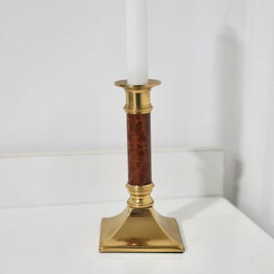 May include: A brass candlestick with a square base and a brown, patterned central column. A white candle sits atop the candlestick. The candlestick is a decorative home accent.