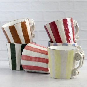 May include: Five ceramic mugs with striped patterns in various colors, including brown, red, green, yellow, and pink. The mugs are stacked and arranged on a white brick background.