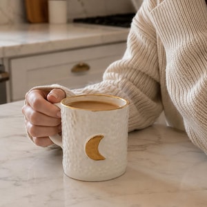 May include: White ceramic mug with a textured finish and a gold crescent moon design. The rim is trimmed with gold. The mug contains a light brown liquid.