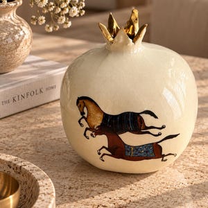 May include: A cream-colored ceramic pomegranate-shaped vessel with a glossy crackle glaze finish. The top features a metallic gold crown-like stem. The body is decorated with a detailed illustration of two galloping horses wearing patterned blankets.