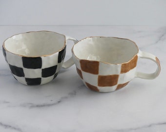 Puede incluir: Dos tazas de cerámica con un patrón a cuadros. Una taza es negra y blanca, y la otra es marrón y blanca. Ambas tazas tienen un borde dorado.
