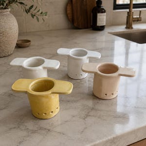 May include: Four ceramic tea infusers in white, yellow, and beige are arranged on a marble countertop. Each infuser has a cylindrical body with a series of small holes and a flat, wing-like handle on either side. The infusers are designed for steeping loose leaf tea.