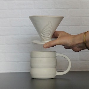 May include: White ceramic coffee dripper with a leaf design and a matching white ceramic mug with a handle.