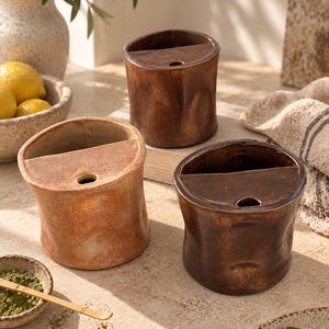 May include: Three cylindrical ceramic vessels with a glossy finish in varying shades of brown. Each container features a flat, semi-circular top section with a small circular opening, creating a distinct divided structure for storage or display.