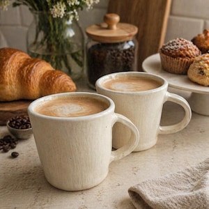 May include: Two off-white ceramic mugs filled with coffee, with a croissant and muffins in the background. A glass jar with a wooden lid and a small bowl of coffee beans are also visible. A linen napkin is in the foreground.
