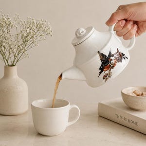 May include: A white ceramic teapot with a floral and bird illustration on the side, featuring a lid with a small skull-shaped knob. The teapot is pouring brown tea into a matching white ceramic mug, with a book titled THIS IS HOME nearby.