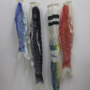 May include: Four koinobori carp streamers hanging from a horizontal wooden rod with black metal clips. The set includes a blue fish, a black fish, a white streamer with colorful patterns, and a red fish, all made of lightweight fabric material.