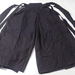 May include: Black hakama pants with long, attached ties. The wide-legged pants are made of a dark fabric and are laid out flat. The ties extend from the waist and are designed to be wrapped and tied.