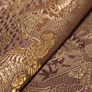 Pode incluir: Close-up de um tecido dobrado com um padrão ornamental e repetitivo. O tecido apresenta uma base roxa escura com desenhos florais e geométricos dourados e bronze. A textura parece detalhada e luxuosa.