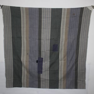 Pode incluir: Têxtil retangular com riscas verticais em tons de cinza, azul e bege. Remendos de tecido azul escuro são costurados no têxtil. O têxtil está pendurado em duas presilhas de metal.