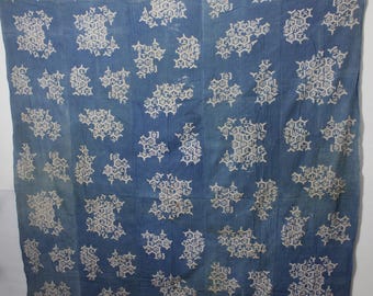 May include: A blue textile with a repeating white floral pattern. The fabric is hanging against a white wall. The pattern is made up of clusters of stylized flowers.