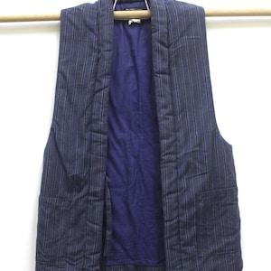 Puede incluir: Un chaleco azul marino con rayas verticales e interior azul oscuro. El chaleco está colgado de una barra de madera sobre un fondo blanco. El chaleco tiene dos bolsillos.