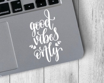 May include: A silver laptop with a white sticker that says "good vibes only" on the top of the trackpad. The laptop is on a white wooden surface.