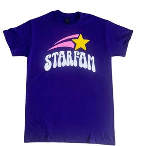 Peut inclure: T-shirt violet avec un graphique rétro blanc d'une étoile filante avec une étoile jaune au sommet et le mot "STARFAM" en dessous.