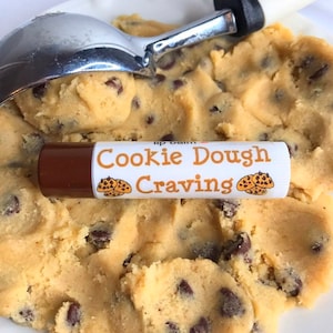 Könnte beinhalten: Ein braunes und weißes Lippenbalsam-Röhrchen mit dem Text "Cookie Dough Craving" und einem Bild von Schokoladen-Chip-Cookies. Der Lippenbalsam liegt auf einer Schüssel mit Keksteig.