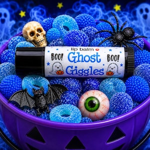 Könnte beinhalten: Ein zylindrischer Lippenbalsam-Stift mit der Aufschrift Ghost Giggles, weißem Hintergrund, blauem Text und Geister-Illustrationen. Er liegt in einem violetten Eimer, gefüllt mit blauen Gummiringen, einem kleinen Totenkopf, einer Spinne und einem Auge.