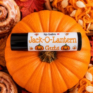 May include: A cylindrical lip balm tube with a white label featuring the orange text "Jack-O-Lantern Guts," two small orange jack-o'-lantern illustrations, and several small orange leaf icons, finished with black caps on both ends.