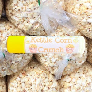 Peut inclure: Un tube de baume à lèvres jaune et blanc avec le texte "Kettle Corn Crunch" et un dessin de pop-corn. Le baume à lèvres est entouré de sacs de pop-corn.