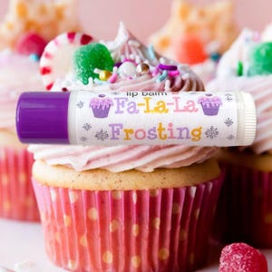 Peut inclure: Un tube de baume à lèvres avec un bouchon violet et l'inscription "Fa-La-La Frosting", décoré de motifs de petits gâteaux et de flocons de neige. Le baume à lèvres repose sur un cupcake avec un glaçage rose et une caissette à pois roses.