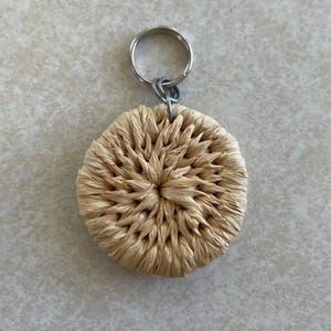 May include: A round, woven raffia keychain with a silver-colored metal keyring. The raffia is a natural beige color, and the weave creates a textured, radial pattern. The keychain is a simple, handmade accessory.