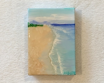 May include: A small painting of a beach scene with a sandy shore, blue water, and a distant mountain range. The painting is signed "Augusta" in the lower right corner.