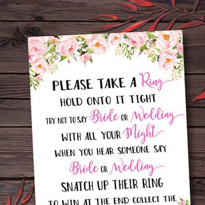May include: A white card with pink floral borders and pink text that reads "PLEASE TAKE A Ring HOLD ONTO IT TIGHT TRY NOT TO SAY Bride OR Wedding WITH ALL YOUR Might WHEN YOU HEAR SOMEONE SAY Bride OR Wedding SNATCH UP THEIR RING TO WIN AT THE END COLLECT THE MOST Bling"