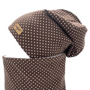 Könnte beinhalten: Eine braune Mütze mit weißen Punkten und ein passender brauner Halsschmuck mit weißen Punkten. Die Mütze hat ein kleines braunes Etikett mit dem Wort "nadiyat" darauf.