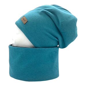 Puede incluir: Conjunto de gorro y braga de cuello color turquesa. El gorro tiene un diseño holgado y una pequeña etiqueta rectangular marrón. La braga de cuello es un tubo ancho de la misma tela turquesa. El conjunto se muestra sobre una cabeza de maniquí blanca.