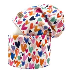 Peut inclure: Ensemble bonnet et tour de cou blancs ornés d'un motif de cœurs colorés roses, oranges, bleus et verts. Le bonnet porte une petite étiquette marron. Le tour de cou est de forme tubulaire.