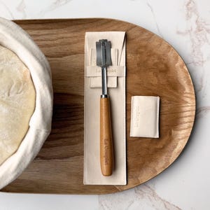 May include: A bread lame with a wooden handle and a metal blade, resting on a cream-colored fabric sleeve. The handle is branded with the text "La Couplet". A small, matching cream-colored fabric pouch with the same text sits beside it.