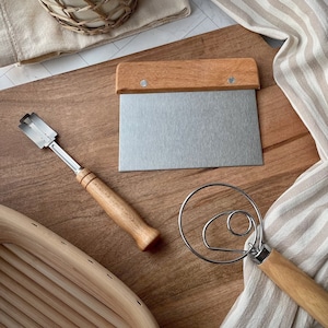 May include: A wooden cutting board with a metal dough scraper, a metal pastry cutter, and a wooden dough whisk. The cutting board is made of light brown wood and has a natural finish. The dough scraper has a wooden handle and a metal blade. The pastry cutter has a wooden handle and a metal blade. The dough whisk has a wooden handle and a metal whisk.