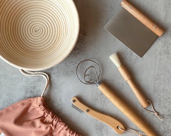 May include: A bread-making kit with a round, coiled proofing basket, a dough scraper, a whisk, a lame, a pastry brush, and a pink drawstring bag. The tools have wooden handles and are arranged on a gray surface. The bag has the text "La Couplet".