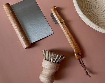 May include: A collection of baking tools, including a stainless steel dough scraper with a wooden handle, a bread lame, and a small brush with a wooden handle. The tools are arranged on a muted red-brown surface. A woven banneton basket is in the background.