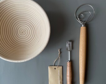 May include: A light-colored wooden banneton proofing basket, a bread lame with a wooden handle, a dough whisk, and a tag that reads "Bread Lame Blades". The items are arranged on a gray surface. The brand name "La Couplet" is at the bottom right.