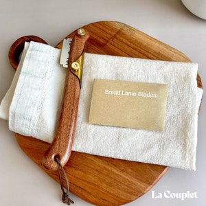 May include: A wooden bread lame with a gold metal blade and a leather strap. The lame is on a wooden cutting board with a white cloth and a brown card that says "Bread Lame Blades".
